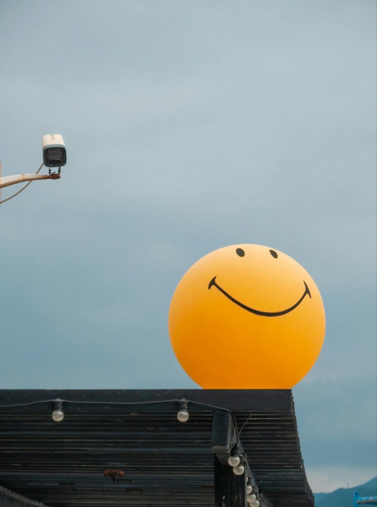 A large yellow smiley face balloon on top of a building, with a cloudy sky in the background.